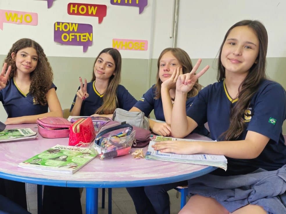 Alunas do Fundamental II sorriem em volta de uma mesa, na sala com cartazes de perguntas em inglês