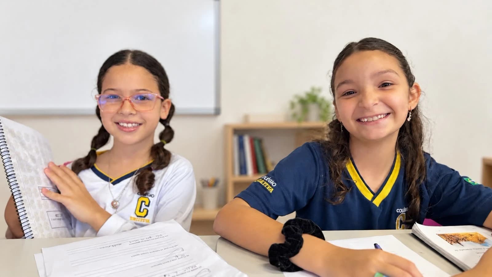 Duas alunas sorriem na sala de aula, com os cadernos abertos sobre a mesa