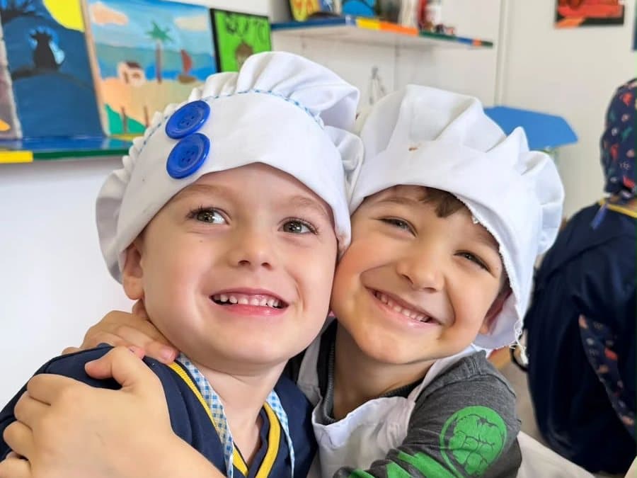 Dois alunos abraçados e sorrindo, de chapéu de chef, na oficina de culinária