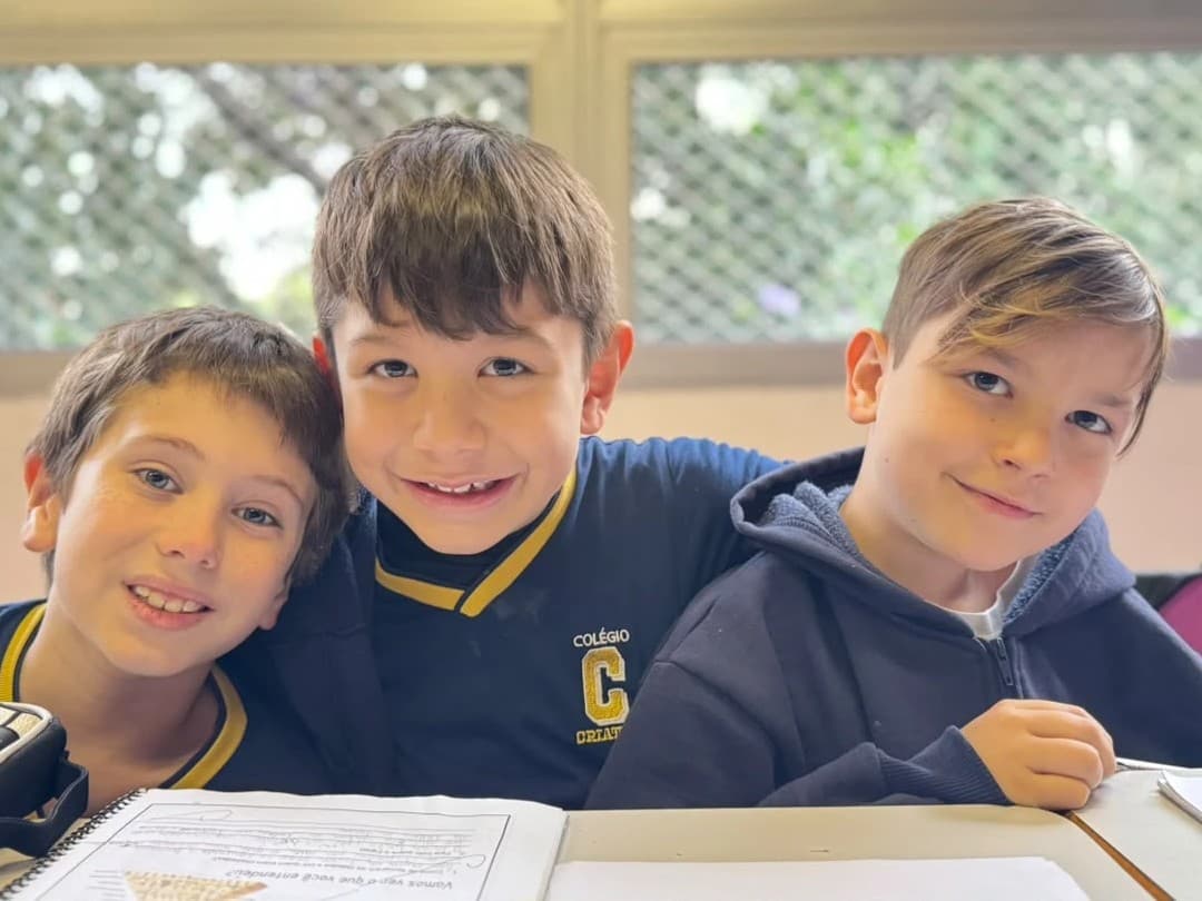 Três alunos dos anos iniciais sorriem abraçados na carteira, diante da janela