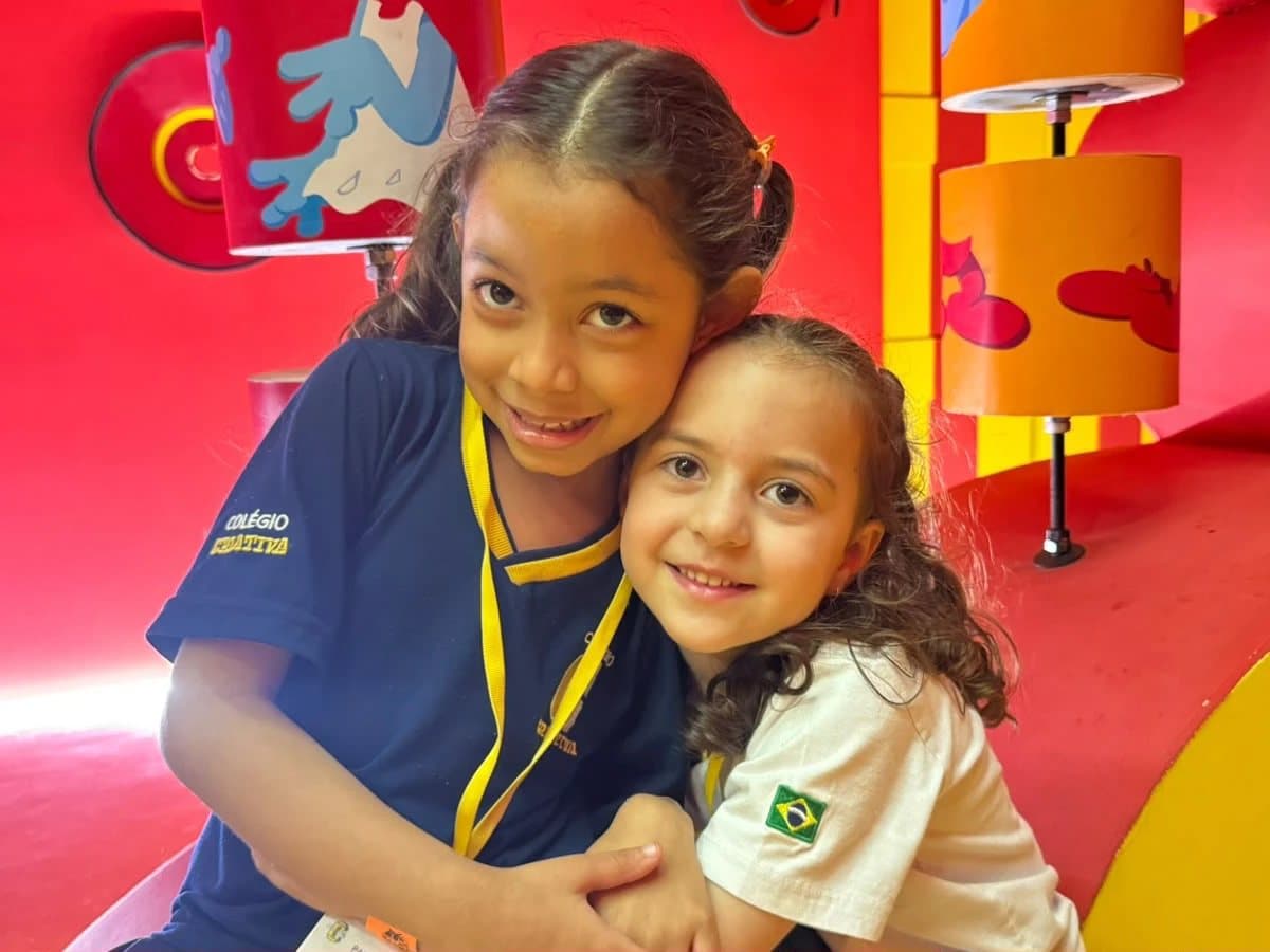 Duas alunas abraçadas e sorrindo diante de uma parede vermelha