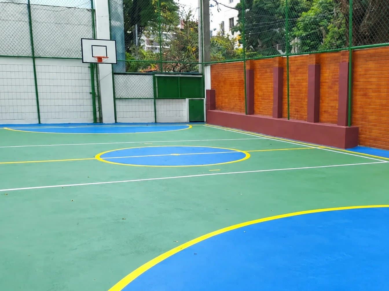 Quadra poliesportiva ao ar livre, com piso verde e azul e tabela de basquete
