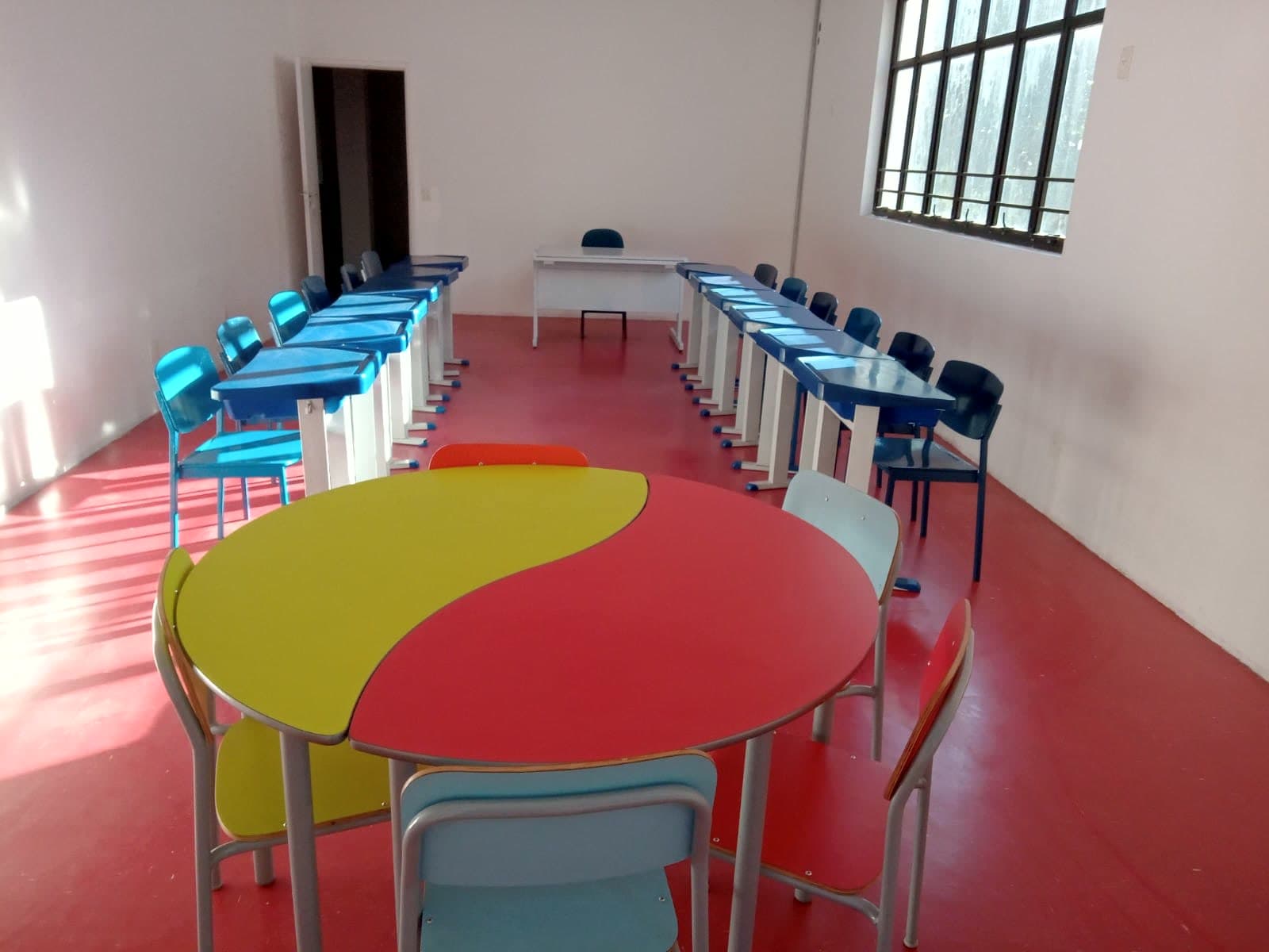Sala ampla e iluminada, com mesas coloridas em amarelo e vermelho e carteiras junto às janelas